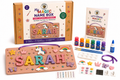 A children's craft kit called 'My Magic Name Box' shown with its packaging box, a completed wooden name box sample, a step-by-step guide, small paint bottles, brushes, ribbon, stickers, and other accessories on a white background.