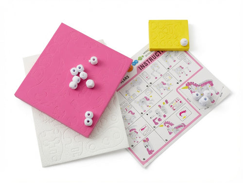 A children's craft or educational toy set on a white background, featuring a bright pink square tile with embossed patterns, a white textured square tile, a yellow square tile, a printed instruction sheet, and several small white googly eyes.